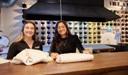 Two team members at the Blarney Aran Experience in Blarney Woollen Mills, Cork, presenting handcrafted Irish Aran sweaters with colourful wool displays in the background