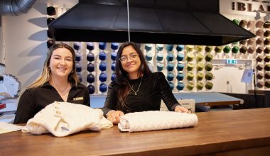 Two team members at the Blarney Aran Experience in Blarney Woollen Mills, Cork, presenting handcrafted Irish Aran sweaters with colourful wool displays in the background