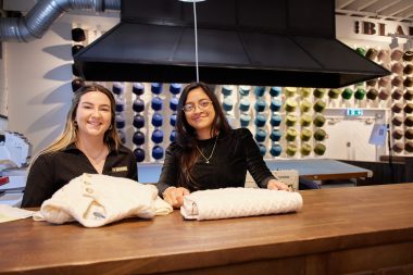 Two team members at the Blarney Aran Experience in Blarney Woollen Mills, Cork, presenting handcrafted Irish Aran sweaters with colourful wool displays in the background