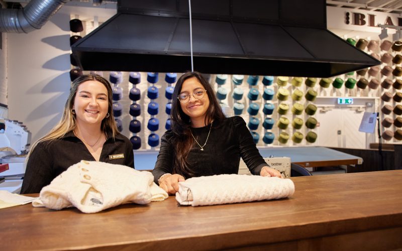 Two team members at the Blarney Aran Experience in Blarney Woollen Mills, Cork, presenting handcrafted Irish Aran sweaters with colourful wool displays in the background
